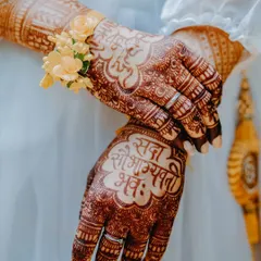 A close-up of the bride's hands, showcasing her intricate mehendi design with the auspicious phrase "Sada Saubhagyavati Bhava". The delicate floral wristlet adds another layer of beauty to this detailed shot.