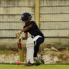 Eyes on the ball. A young cricketer, fully equipped with pads and helmet, is captured mid-shot during a practice session in the nets, demonstrating proper form and concentration.