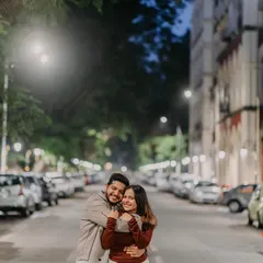 A cozy, candid hug on a quiet, rain-slicked street in Mumbai. The soft streetlights and classic architecture create a warm, romantic atmosphere for this pre-wedding moment.