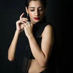 An elegant low-key portrait of a woman in a black saree. The lighting gently illuminates her face and hands, highlighting her red lipstick and diamond jewelry against the dark background.