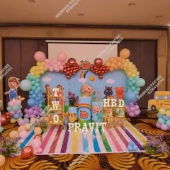 An elaborate Cocomelon stage decoration for a second birthday, complete with a rainbow balloon arch, character props, and a striped floor mat.