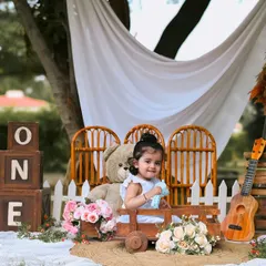 A wide shot of the whimsical outdoor setup for a first birthday. The scene includes "ONE" blocks, a teddy bear, a ukulele, and floral arrangements, creating a perfect boho-inspired setting for her milestone photos.