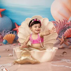 A whimsical and creative baby photoshoot featuring a little one as an adorable mermaid, waving from inside a giant seashell on a beach-themed set.