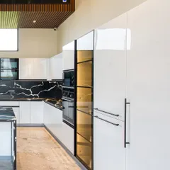 A modern, open-plan kitchen with high-gloss white cabinets and a bold black marble backsplash and island. The wood-paneled ceiling detail adds warmth and defines the kitchen space.
