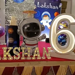 A close-up of the personalized details for Lakshan's space party. The setup includes a large astronaut cutout, a light-up number '6' with a planet inside, and illuminated letters for the name.