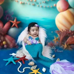 A fun and creative mermaid-themed photoshoot for a baby girl. The setup includes a knitted mermaid tail, seashell props, and an underwater-themed backdrop to create a magical scene.
