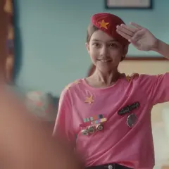 A close-up of the young character saluting her reflection. The combination of the red beret and the imaginative, self-made uniform was designed to show her early determination and the start of her journey.