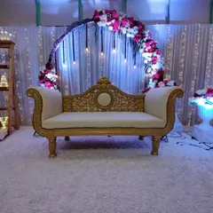 This elegant baby shower stage is available for rent. The setup includes a beautiful golden sofa, a semi-circular floral arch with hanging Edison bulbs, and "BABY" marquee letters on a wooden ladder shelf.