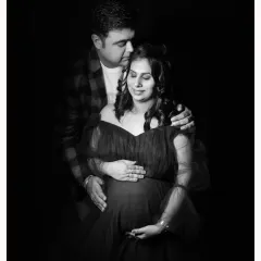 An intimate black and white studio portrait of a couple. The partner's hands gently rest on the mother-to-be's shoulders and bump.