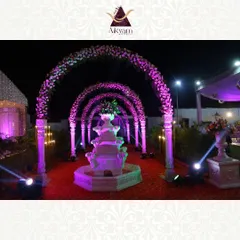 A grand entrance walkway featuring a series of floral arches over a red carpet, leading to a beautiful water fountain. The purple lighting creates a dramatic and luxurious welcome for guests.