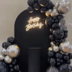 A sleek and modern birthday party backdrop. This setup features a black arched panel with a "Happy Birthday" neon sign, framed by a sophisticated arrangement of black and silver balloons.