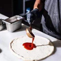 The first layer of flavor. I use a simple, rich San Marzano tomato sauce, spreading it carefully to leave room for that perfect, puffy crust to rise in the oven. It's the sweet and tangy base for all our classic pizzas.