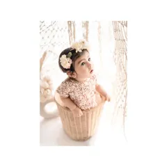 A baby girl in a sequined dress peeks out of a wicker basket, looking up with wonder. The clean, white background and bohemian-style props create a chic and adorable setup for a milestone photoshoot.