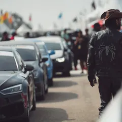 A participant in a custom leather jacket walks through a lineup of performance cars, soaking in the atmosphere of the auto expo.