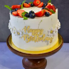 A beautiful birthday cake topped with a crown of fresh berries. This simple yet elegant design features fresh strawberries and blueberries, delicate white daisies, and a gold "Happy Birthday" script.