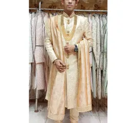 A full-length view of a groom in a light beige silk sherwani. The outfit is accessorized with a gold mala, showcasing a complete and elegant look available for rent.