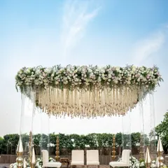 A closer look at the floating mandap, highlighting the dense arrangement of white and pastel pink flowers that form the canopy. The design is complemented by traditional brass elements on the stage.