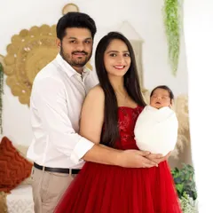 A classic family portrait with a newborn. The proud parents are smiling, holding their swaddled baby, in a warm, boho-chic studio setting.