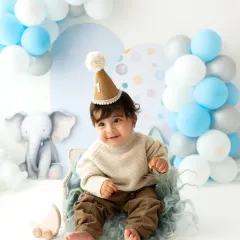 A baby boy wearing a cute party hat for his first birthday photoshoot. The elephant and balloon theme creates a sweet and celebratory atmosphere.