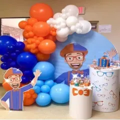 An energetic Blippi themed birthday party with his signature orange and blue colors. The setup includes a large character cutout, a themed backdrop, and custom pedestals decorated with glasses and bowties.