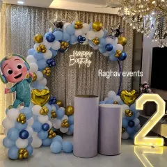 A Cocomelon themed second birthday party decoration. The setup features a balloon arch with a Cocomelon character, a neon sign, cake stands, and a large light-up number '2'.
