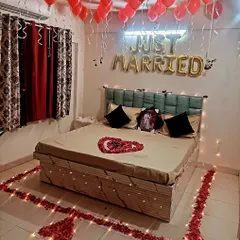 A romantic first-night decoration for a newly married couple, featuring 'Just Married' balloons, a heart made of rose petals on the bed, and a floor border of petals and lights.