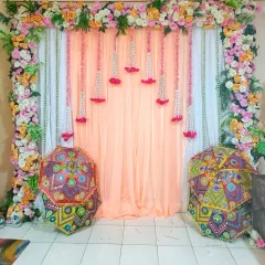 A beautiful backdrop for an at-home Haldi or Mehandi function. The setup includes a floral frame, hanging flower strings, and traditional Rajasthani umbrellas for a pop of color.