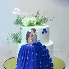 A beautiful two-tier engagement cake with a custom photo topper. The highlight is the bride's flowing blue gown, created with intricate ruffles, and delicate baby's breath flowers.