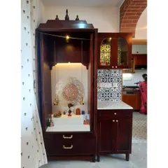This elegant pooja unit is designed to blend seamlessly with the adjacent kitchen cabinetry. It features a tall, open mandir with backlighting and hanging brass lamps, along with an attached storage unit with patterned tiles for a modern touch.