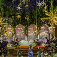 A magical sangeet night with a 'Lilac & Gold' theme. The ceiling is a canopy of lush greenery and hanging stars, with crystal chandeliers casting a warm glow over the entire space.