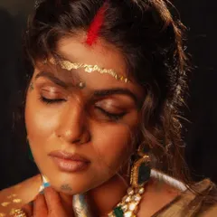 A close-up on the artistic makeup and adornments. The red feather, gold leaf on the forehead, and raw jewelry are all part of the conceptual styling to create a non-traditional, powerful look.