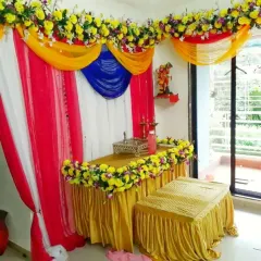 A simple and colorful Ganpati pooja corner for a home. This setup uses bright fabric drapes in yellow, pink, and blue, with yellow floral arrangements on the table and as a top border.