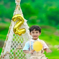A happy birthday boy holds a yellow balloon, sitting by a macrame teepee during his outdoor second birthday celebration.