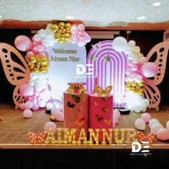 A modern and chic butterfly-themed welcome party for Aiman Nur. The design includes large butterfly wing props, an arched backdrop, and a stylish balloon garland in pink, white, and gold.