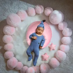 Drifting off to dreamland. This newborn is dressed in a cozy blue sleeper, resting on a pink cushion surrounded by a circle of soft, cloud-like pom-poms. This setup highlights the peaceful and serene nature of a sleeping baby.