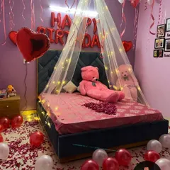 A cozy and romantic birthday room surprise. The setup features a bed with a light-up canopy, "HAPPY BIRTHDAY" foil balloons, a large teddy bear, and the floor covered in red and white balloons with rose petals.