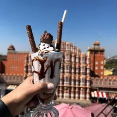 A decadent Ferrero Rocher shake held up against the Hawa Mahal. This shot is perfect for social media, combining a drool-worthy dessert with a world-famous landmark for a highly shareable image.