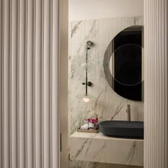 Inspired by Parisian style, this powder room exudes luxury and refinement. A peek through the fluted doorway reveals a space where rich marble textures, a dark stone basin, and warm ambient lighting create a lavish and elegant ambiance.
