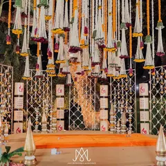 A magnificent stage setup for a traditional ceremony, featuring a backdrop of 'Shubh' motifs, brass elements, and a stunning ceiling of hanging floral tassels.