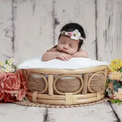 A baby girl with a floral headband resting in a beautiful wicker basket, surrounded by bouquets of pink and yellow flowers.
