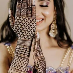 A joyful bride peeking through her intricately designed hands. The design features a mix of geometric jaali work and delicate heart-shaped patterns on the arms.