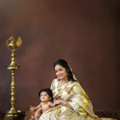 A stunning portrait of a mother and her baby in a traditional 'Mallu' themed setup. The rich brown backdrop and the classic brass lamp (Nilavilakku) add to the authenticity and warmth.