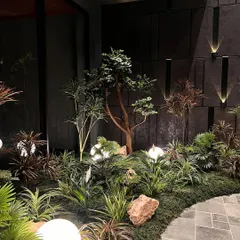 A wide view of an indoor garden featuring a variety of artificial plants, from small bonsai to taller, leafy specimens. The glowing globe lights and mossy ground cover create a complete, immersive landscape.