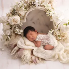 A newborn in a delicate white lace outfit sleeps inside a heart-shaped box filled with white flowers, creating a purely angelic scene.