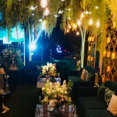 An enchanted forest theme for a reception, with lush greenery hanging overhead, warm Edison bulbs, and plush emerald velvet sofas. This design creates an intimate and luxurious lounge space for guests.