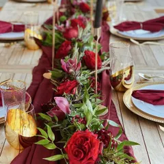 A luxurious and romantic tablescape featuring a deep red color palette. The table is adorned with a lush floral runner of red roses, elegant gold-rimmed plates, and tall black candles.