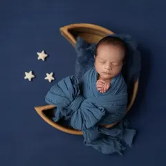 A newborn boy wrapped in a deep blue swaddle, resting in the crescent of a wooden moon prop against a matching blue background. The tiny stars complete this celestial theme.