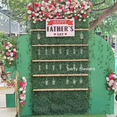 A creative "Cheers to Dad" wall for a Father's Day celebration. The green hedge backdrop is set up to hold drink glasses for guests, topped with a floral arrangement and a "Happy Father's Day" sign.
