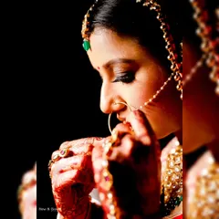 A dramatic and beautiful bridal portrait. The bride is adjusting her nose ring against a pure black background, with warm light illuminating her intricate jewellery and henna.