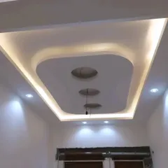 A modern false ceiling design with a central recessed section and curved edges. The combination of indirect cove lighting and spotlights provides balanced illumination.